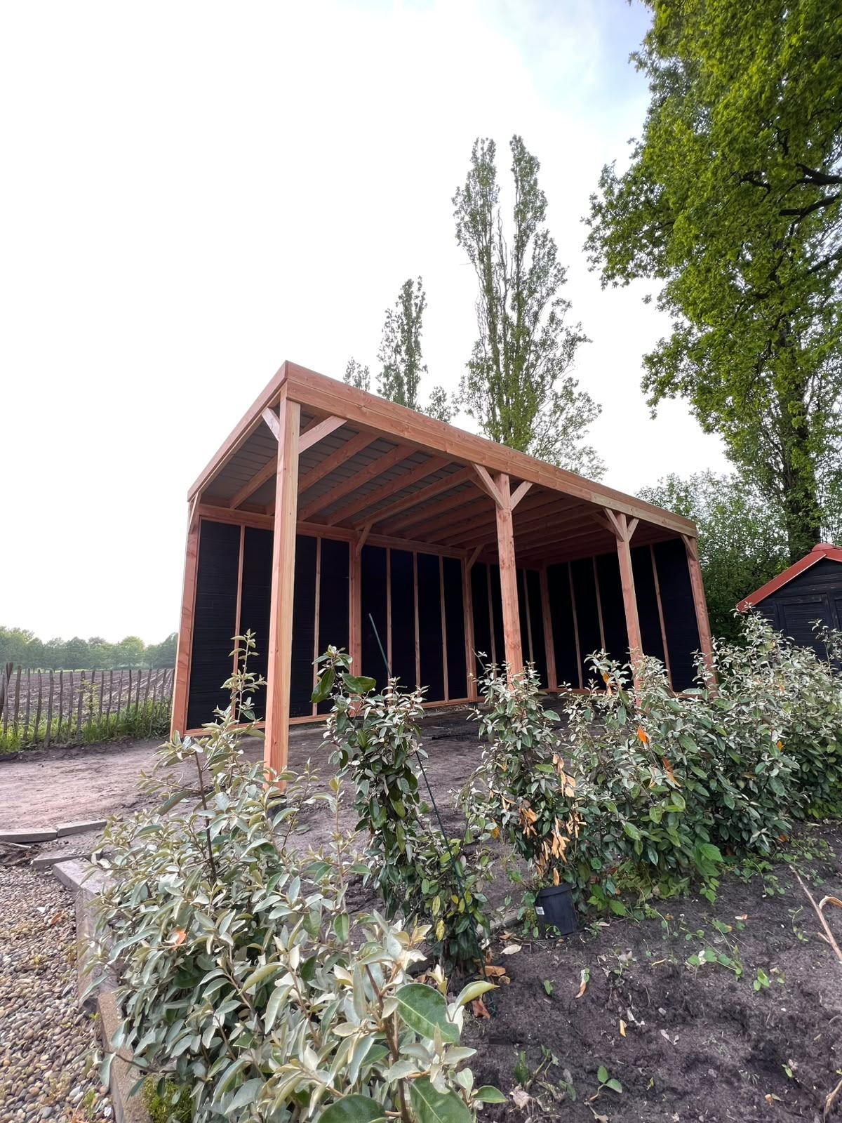 Open houten carport met plat dak aan de rand van een tuin