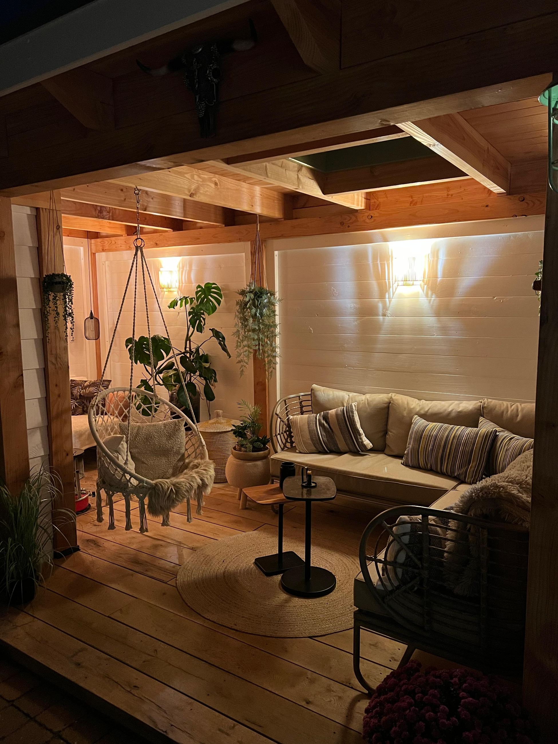 Verlichte overdekte zithoek met hangstoel, loungebank en planten in de avondschemering