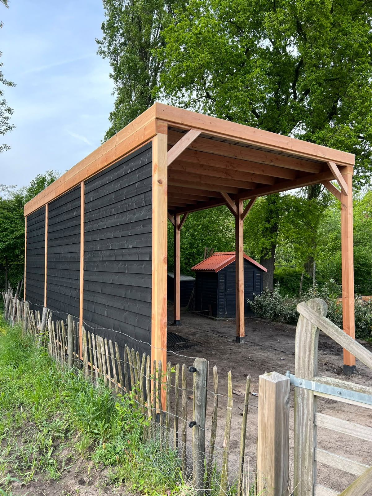 Langgerekte schuilstal met zwarte wand langs een kastanjehouten hek
